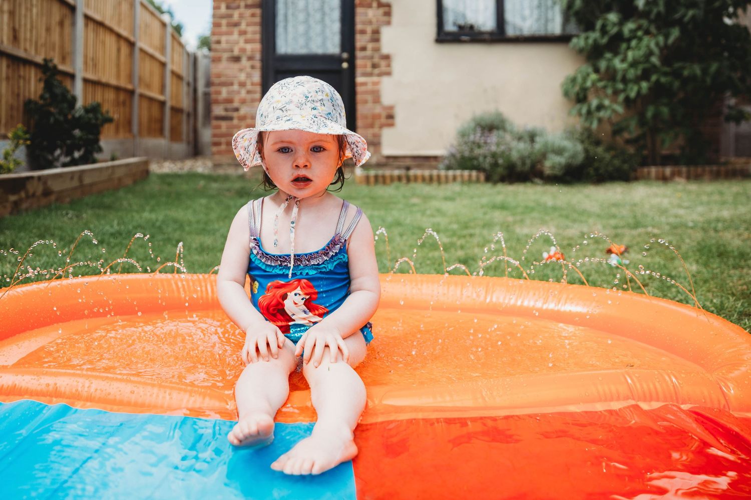 tiny girl sitting in paddling pool wearing sun hat swimming costume