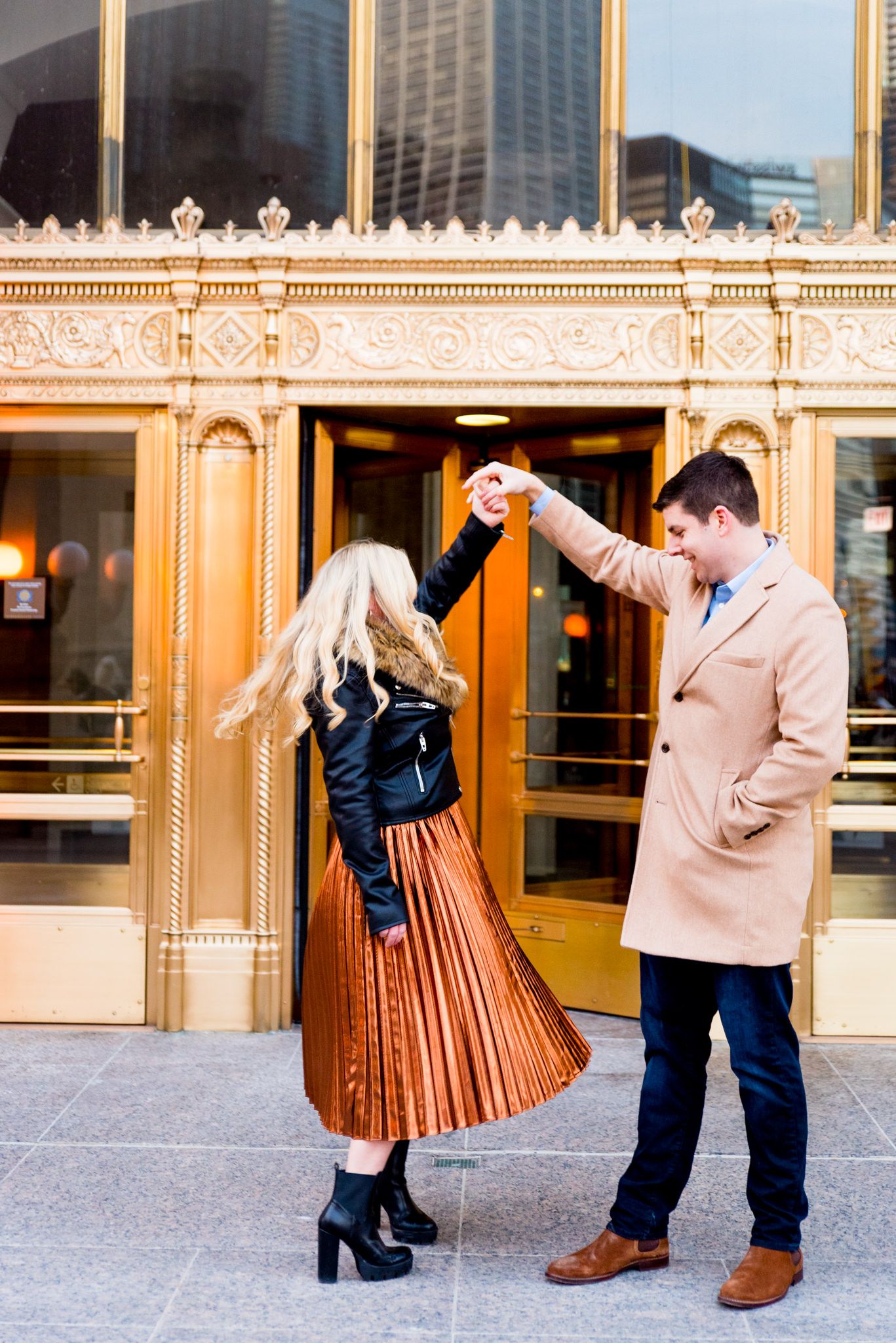 man in tan coat twirling woman in black coat and copper skirt in front of gold doors at Wrigley Building engagement