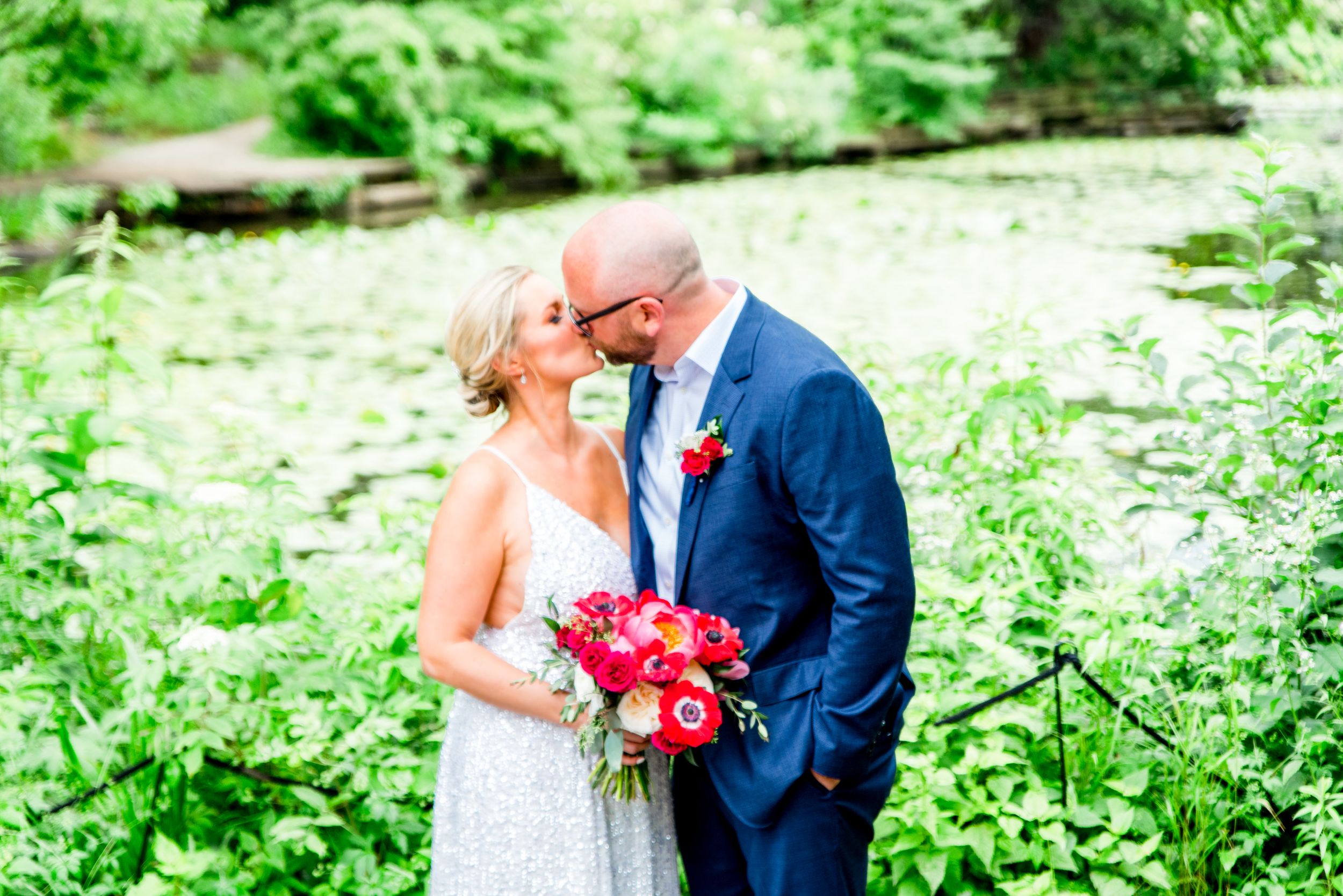 man in navy suit kissing blonde woman in silver dress with red bouquet at Alfred Caldwell Lily Pool in Lincoln Park