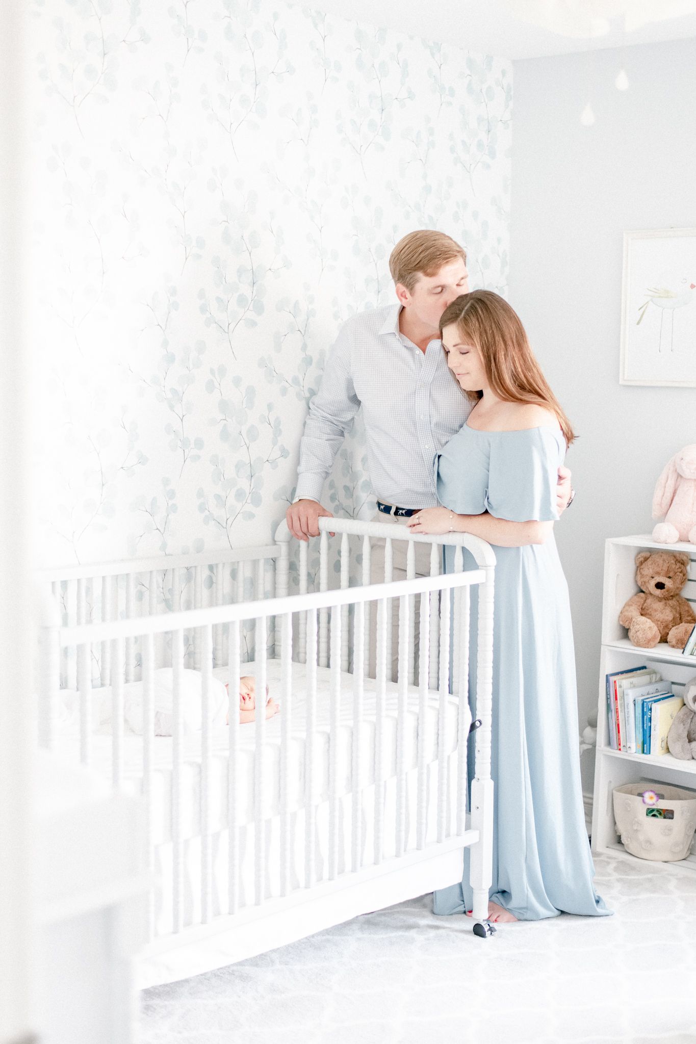 Husband kisses his wife on the head as she gazes at their newborn baby laying in the crib. Mom is wear a blue maxi dress