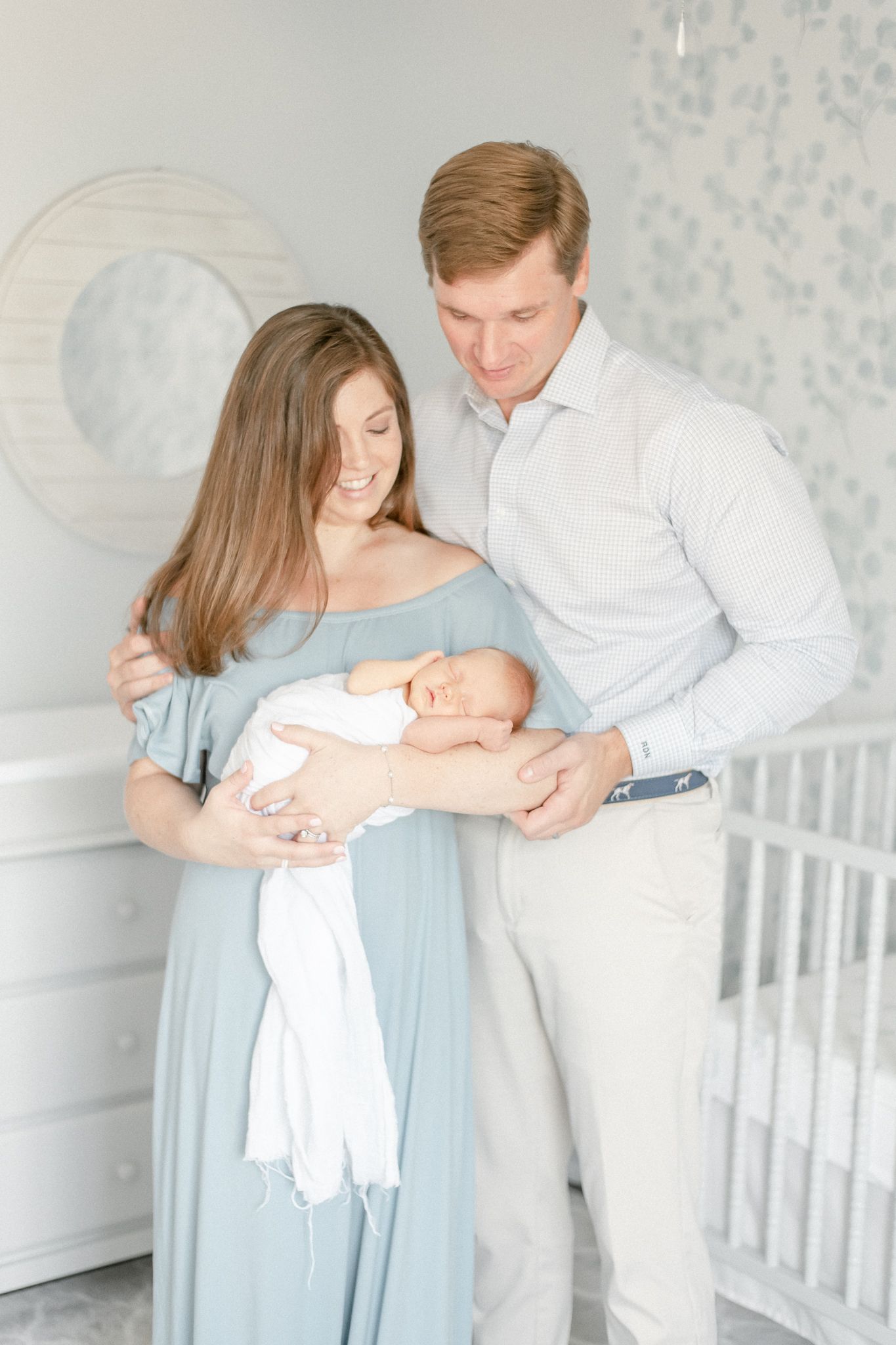 New parents gaze down at newborn as they stand in the blue nursery
