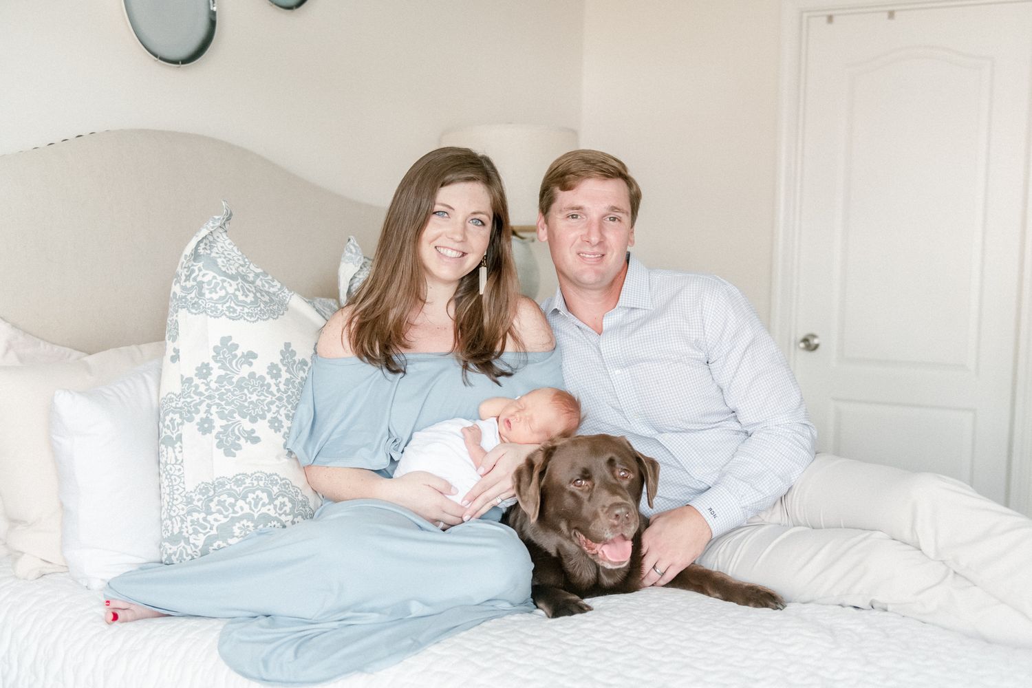 Family holds newborn baby and chocolate lab on their master bed