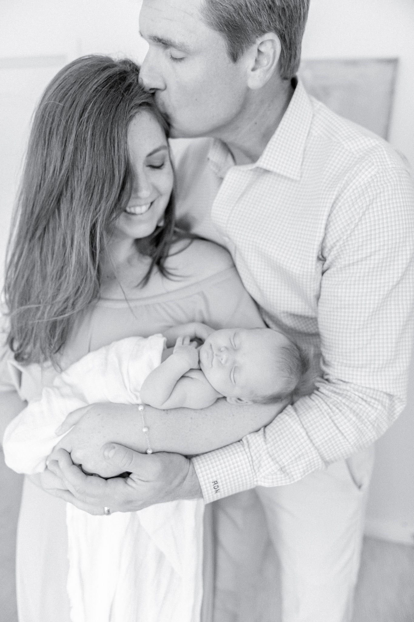 Black and white image of new parents holding newborn, father kisses mother on head