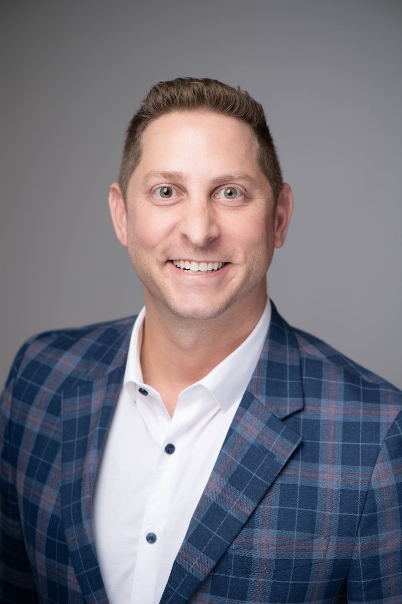Professional headshot of person wearing blue plaid blazer and white shirt against gray background.