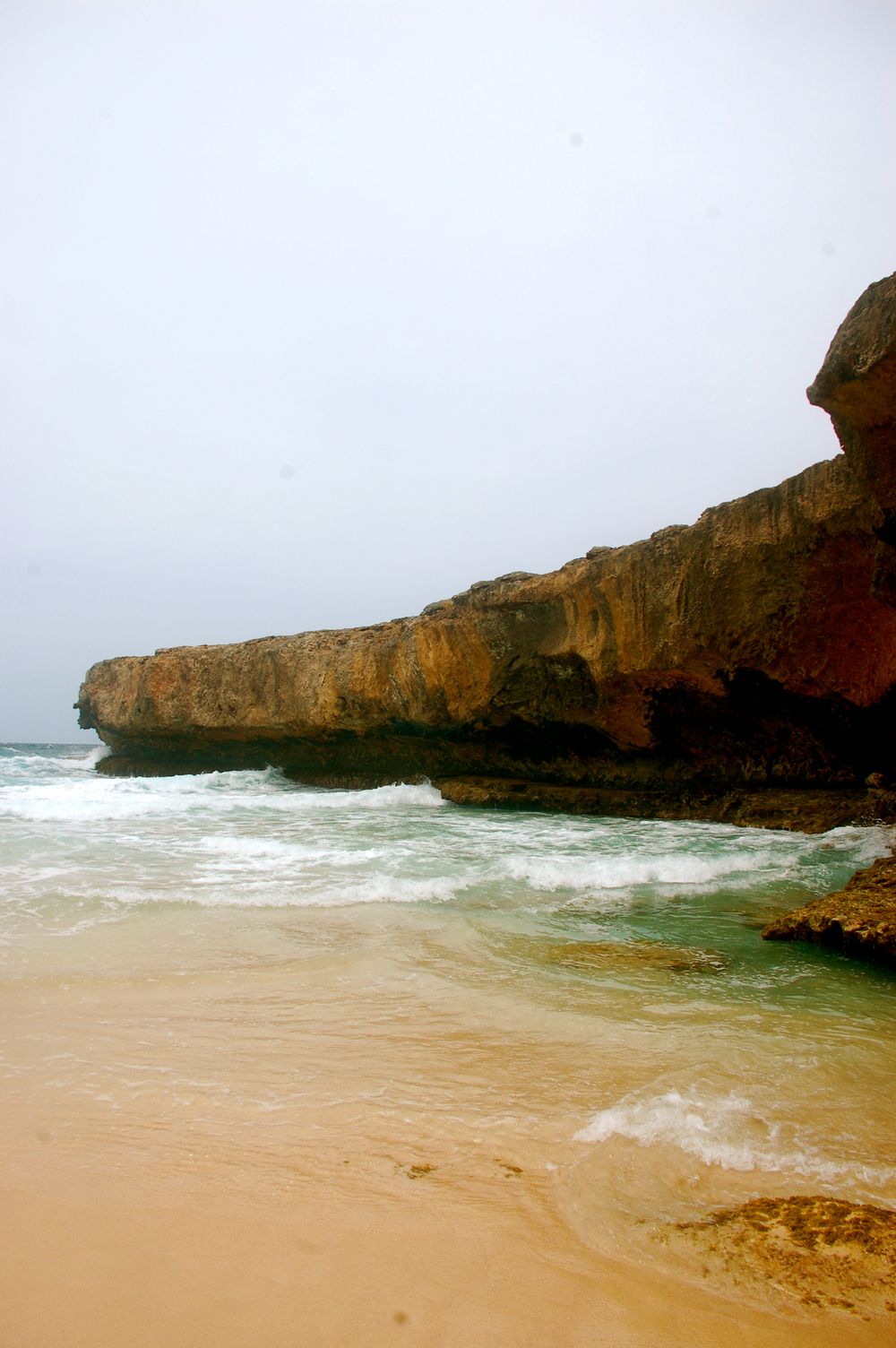 Dramatic coastal cliff meets sandy beach with turquoise waves crashing against rocky shore.