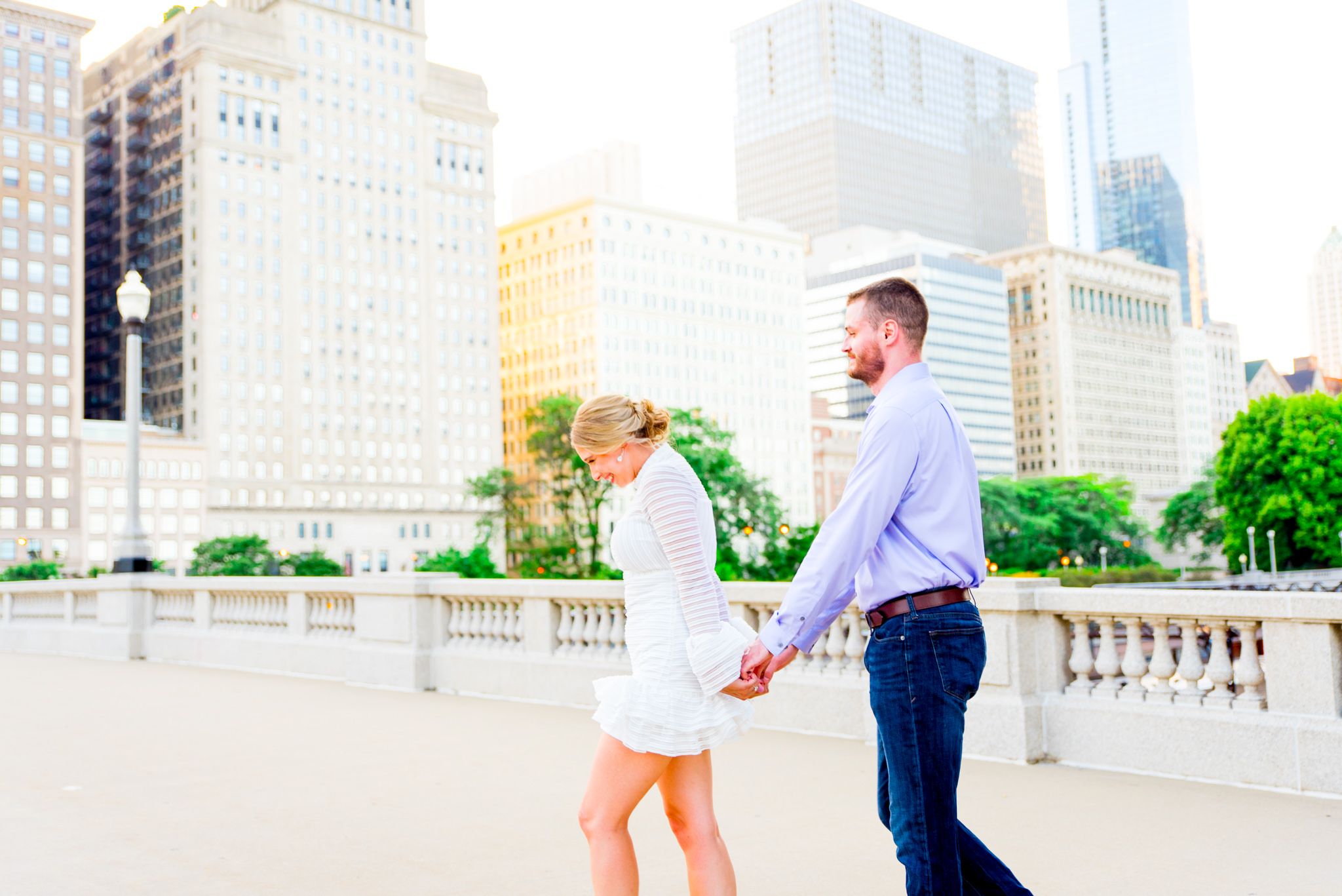 woman in white dress leads man in front of Chicago skyline at sunset engagement pictures