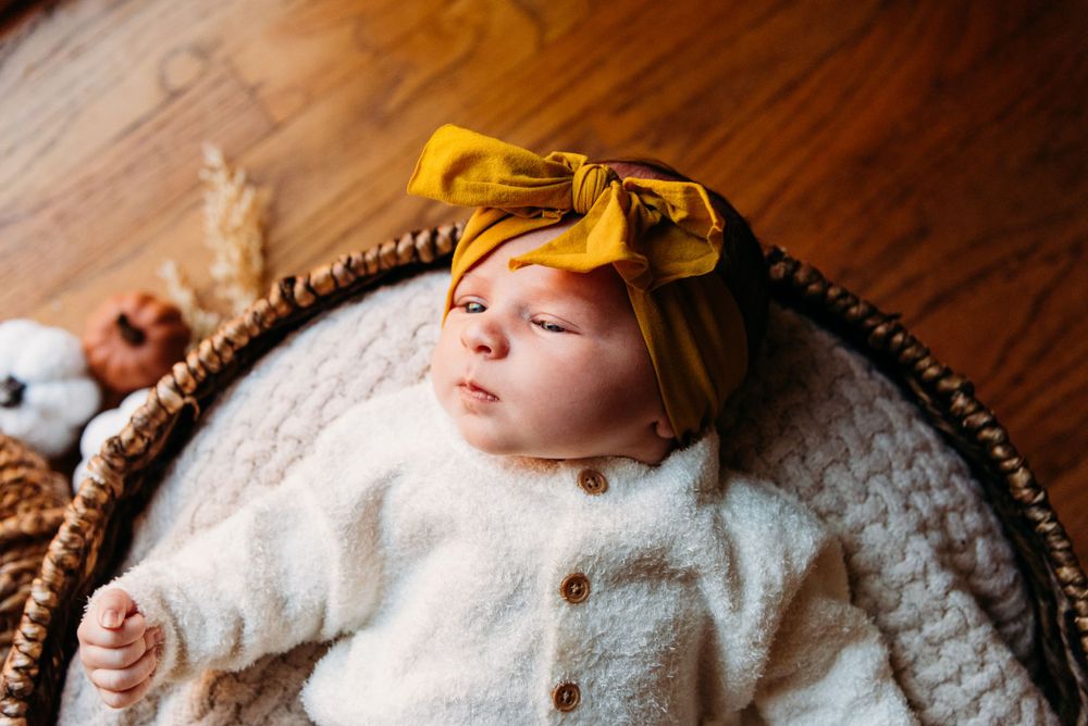 Sleeping baby wearing white sweater and yellow bow headband in basket.