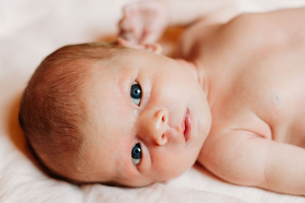 A close-up portrait of a newborn baby with bright blue eyes lying on white bedding.