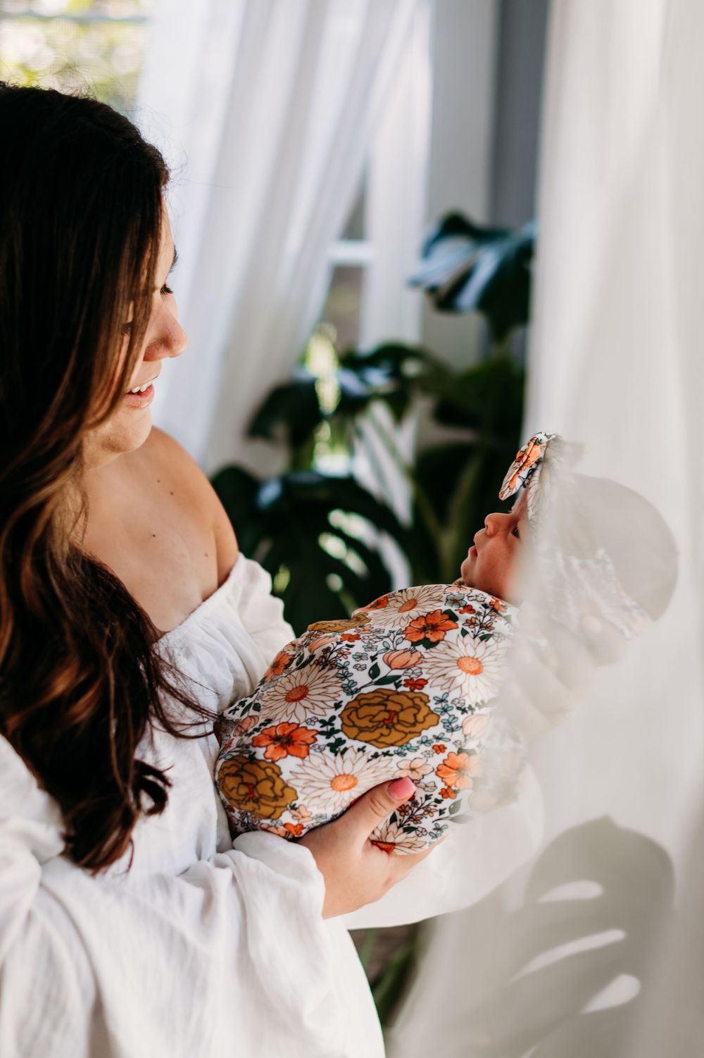 A tender moment between a person in white and a swaddled baby against a bright window with houseplants.