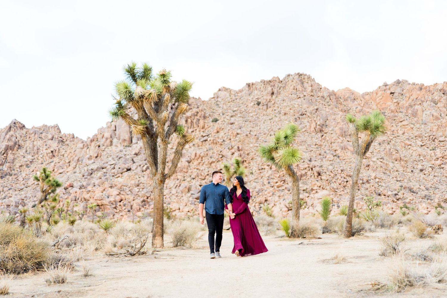 woman in red dress and man in blue shirt smiling and walking through Joshua Tree National Park for engagement pictures