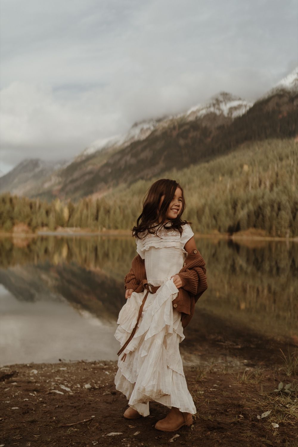 Little girl wearing long white dress and brown sweater standing at Gold Creek Pond in Snoqualmie