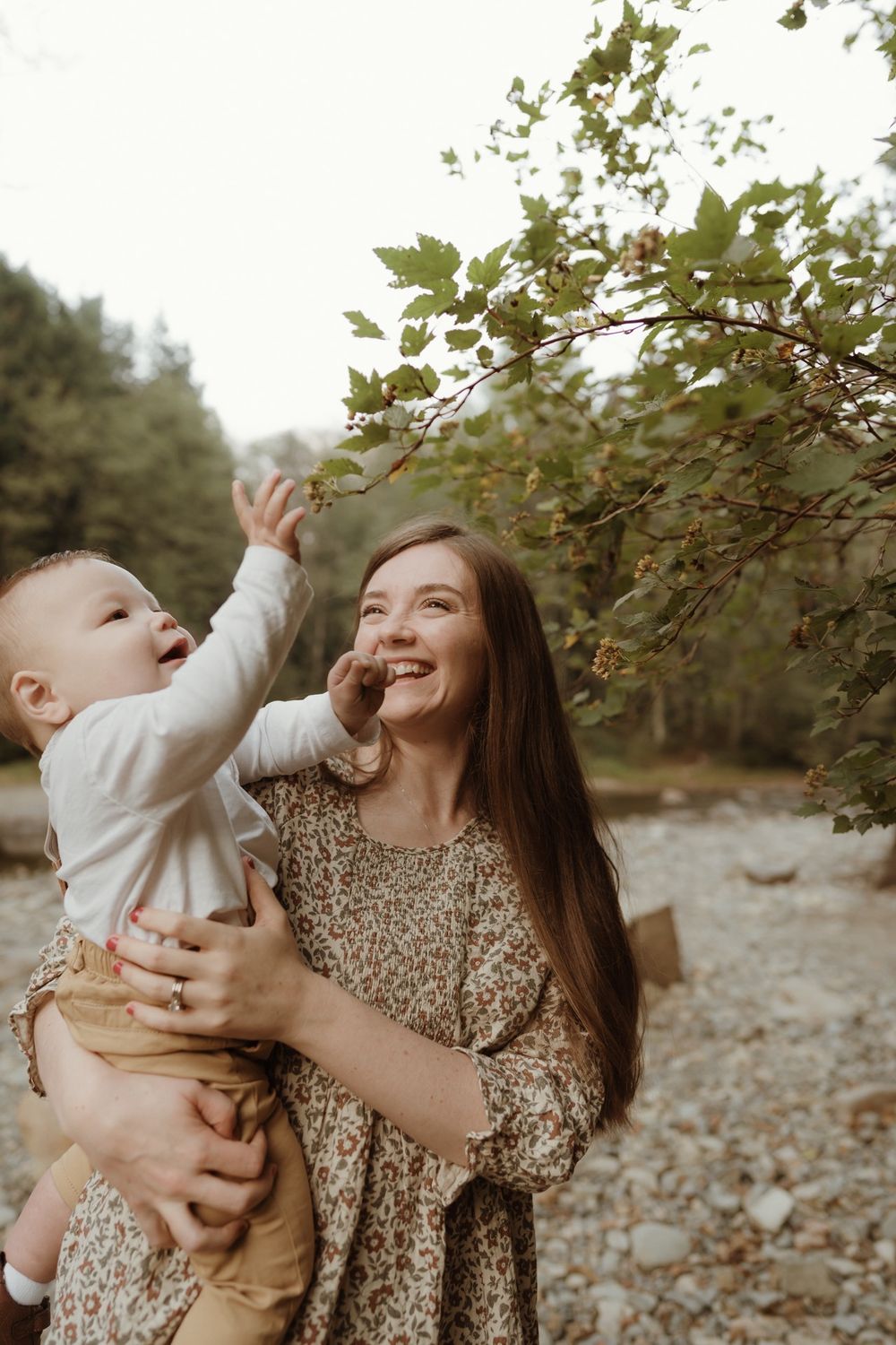 Mother holding up baby boy wearing suspenders reaching for green tree leaves