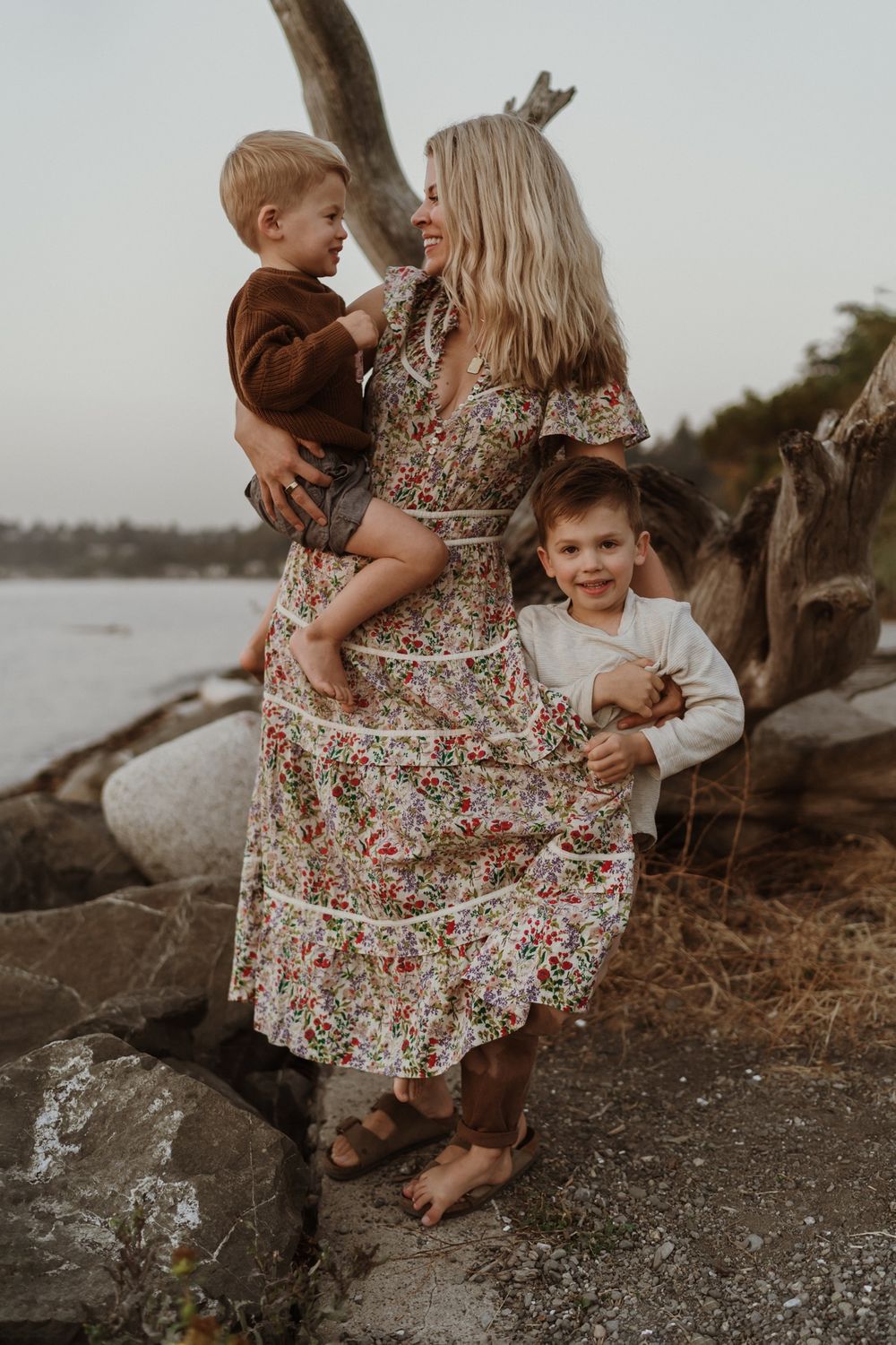 Mother wearing neutral floral dress holding holding boys with driftwood background