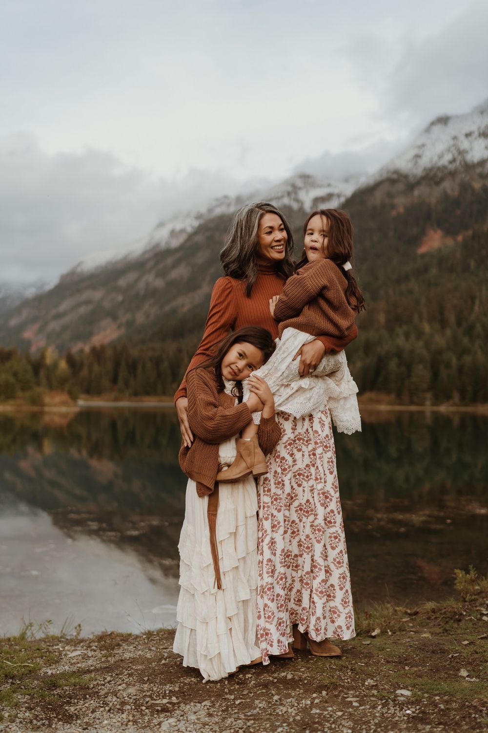 Mother wearing long patterned skirt standing at Gold Creek Pond with daughters wearing white dresses and brown sweaters