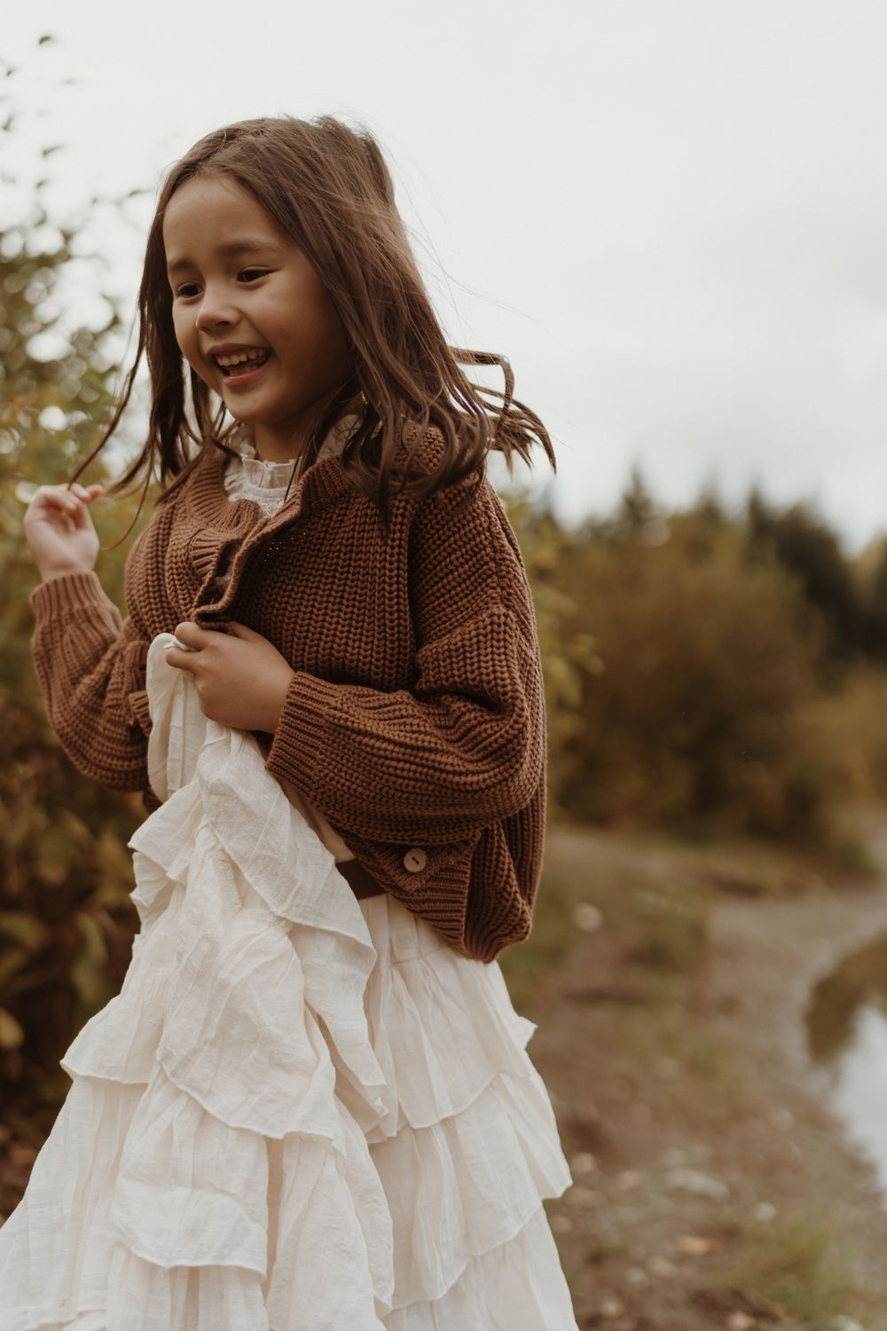 Little girl wearing white dress and brown sweater running along lake