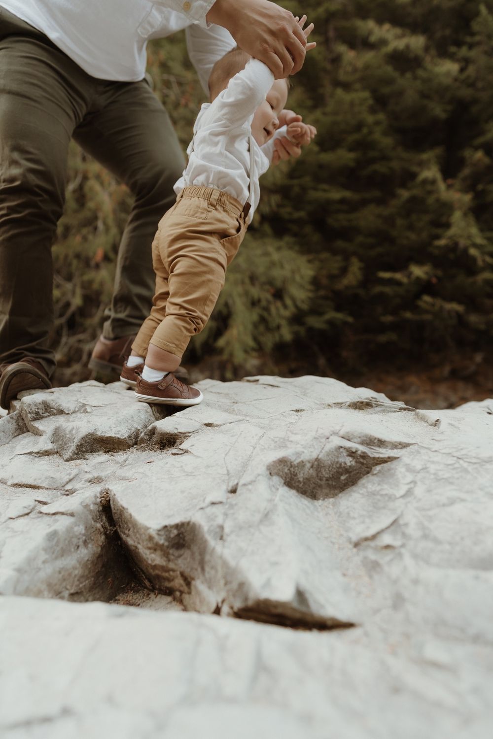 Father holding baby boy wearing yellow pants trying to walk on rock