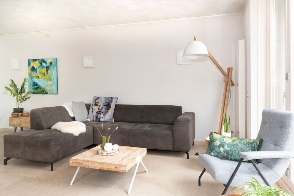Lichte woonkamer met grijze hoekbank, lichtgrijze fauteuil, houten accenten en blauw-gele accessoires