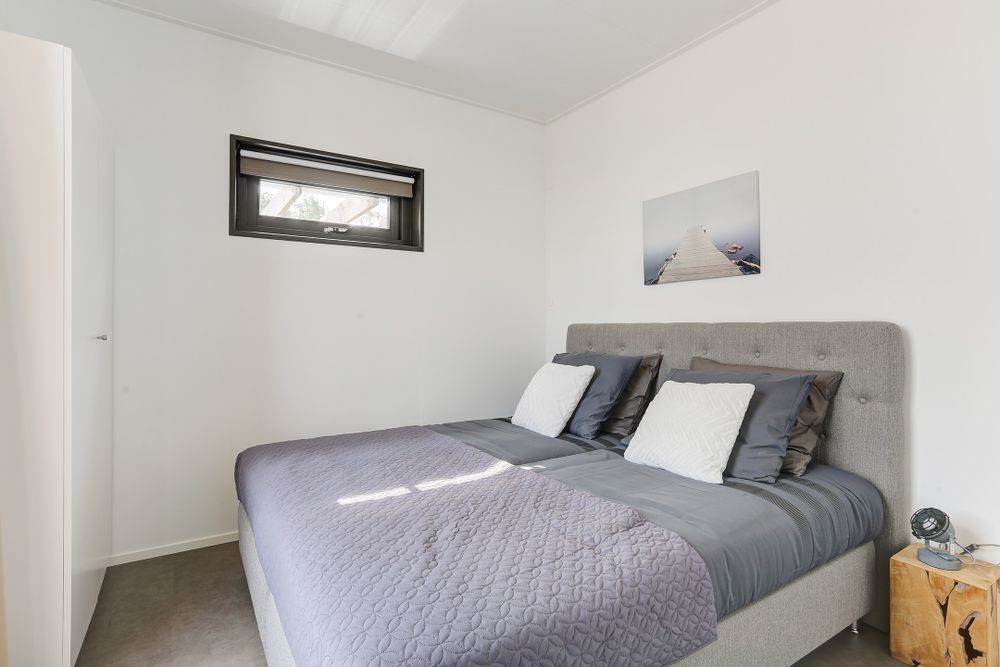 Lichtgrijze boxpring met zes kussens en sprei, een houten bijzettafeltje en foto aan de muur