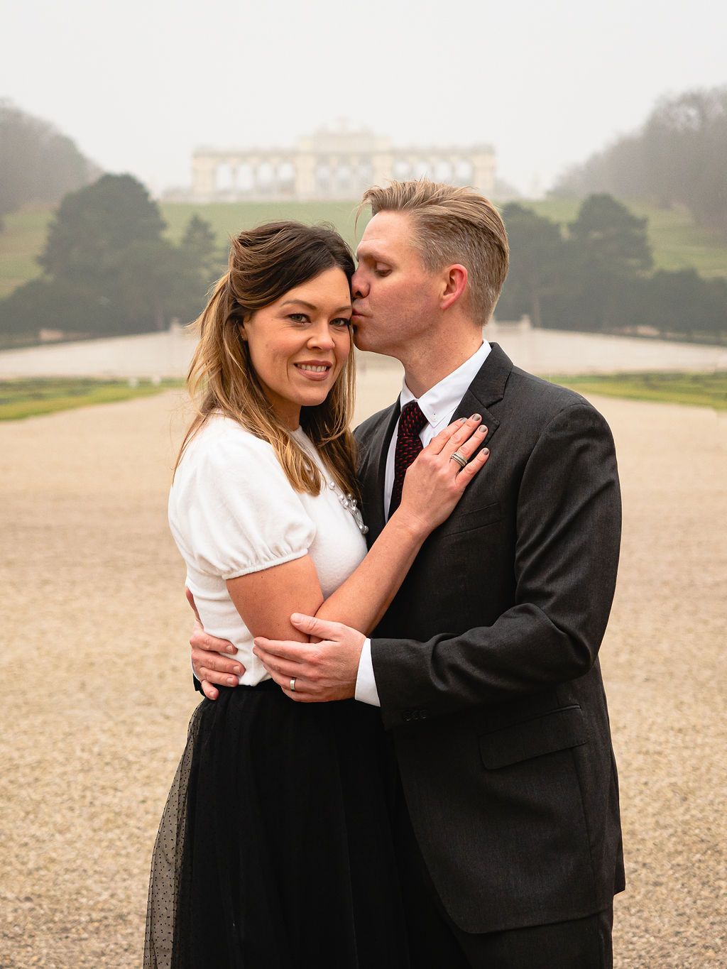 Woman looking at the camera and man is kissing her forehead. Background is the Gloriette in Vienna, Austria