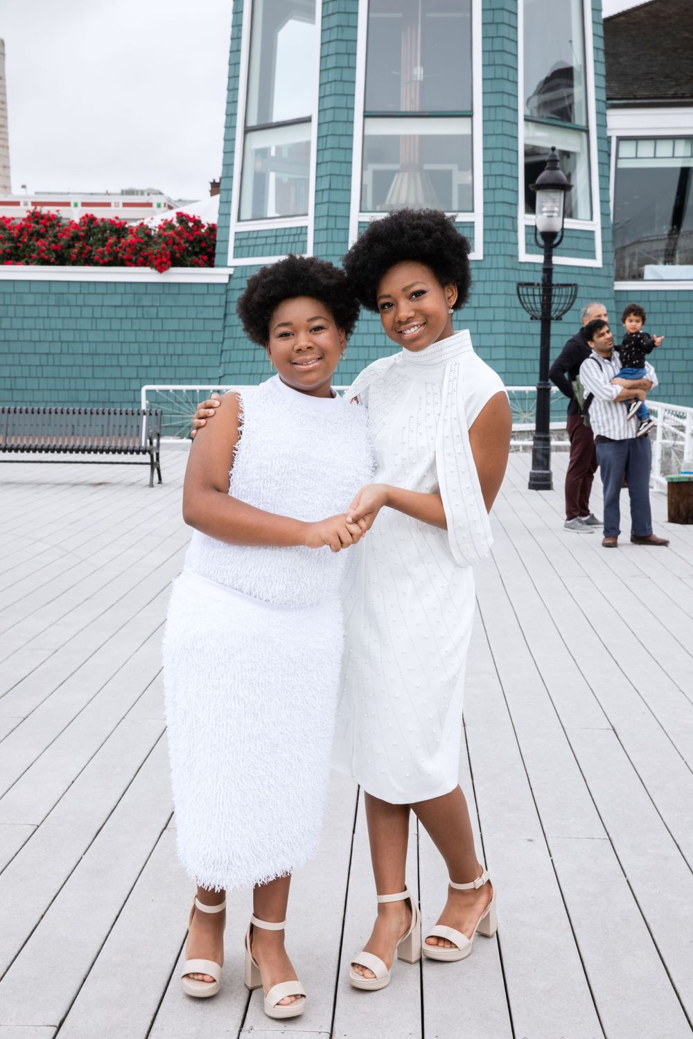 Sisters in matching white dresses pose together on wooden deck with turquoise building behind.