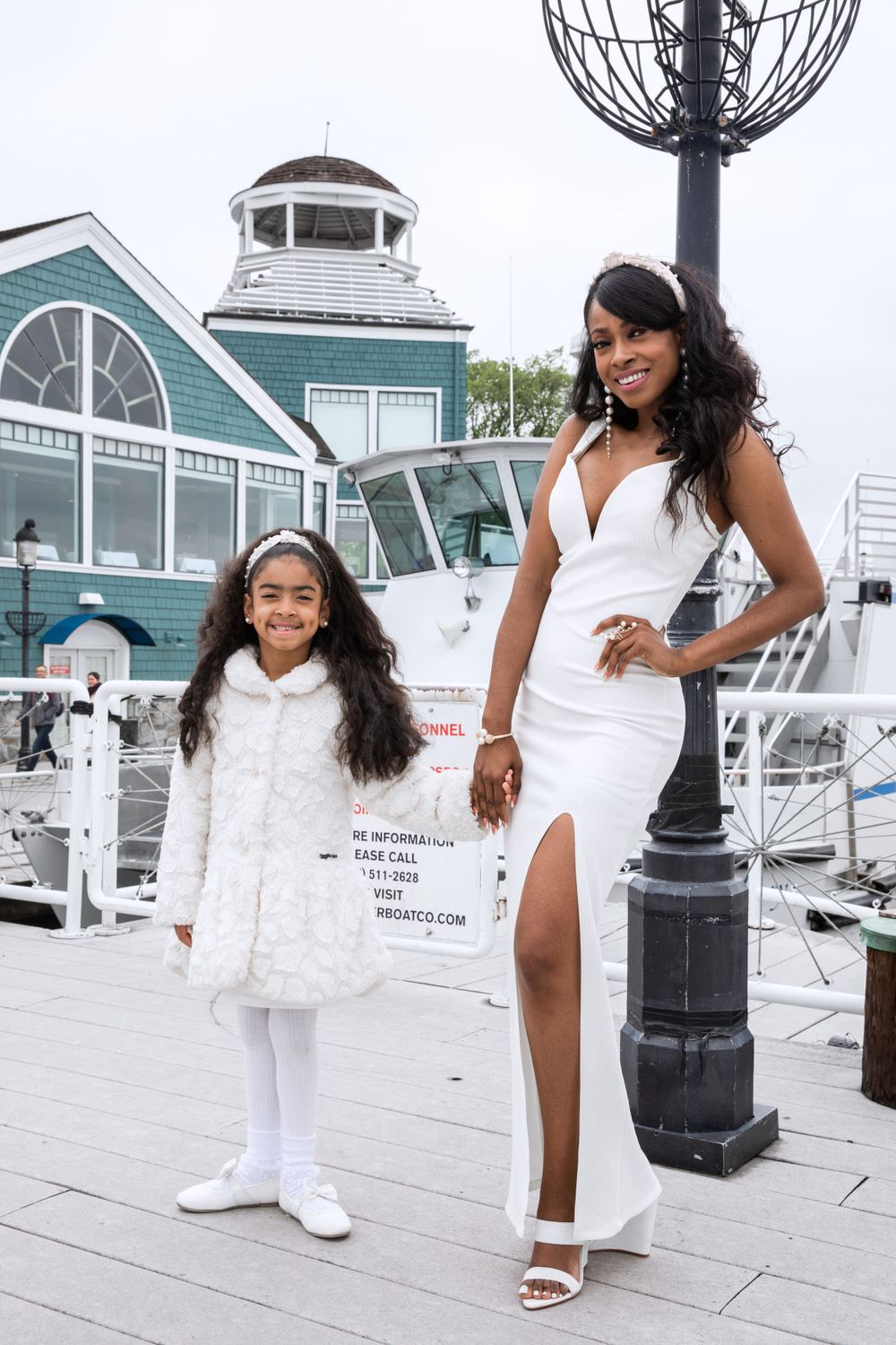 Mom and daughter in elegant white outfits stand together at a marina with boats and buildings in background.