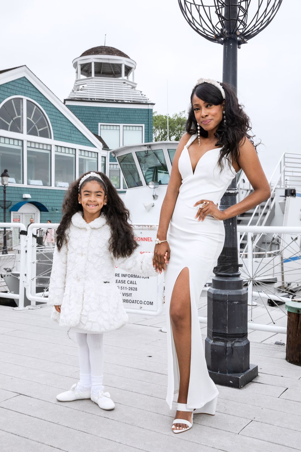 A parent and child wearing coordinated white outfits pose by a marina and lighthouse.