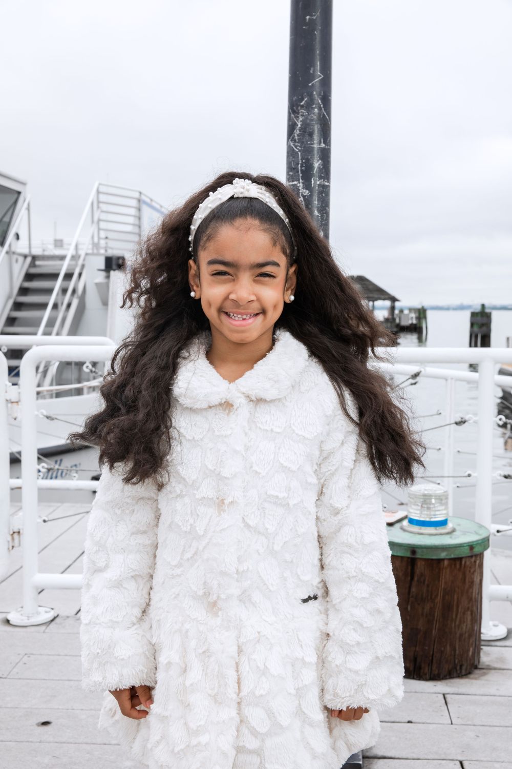 A young person in a white winter coat smiles while standing on a dock.