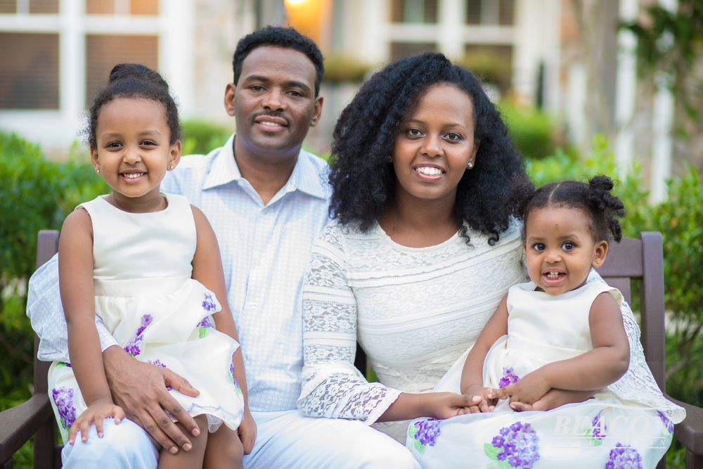 A family of four wearing coordinated white outfits sits together for a portrait outdoors.
