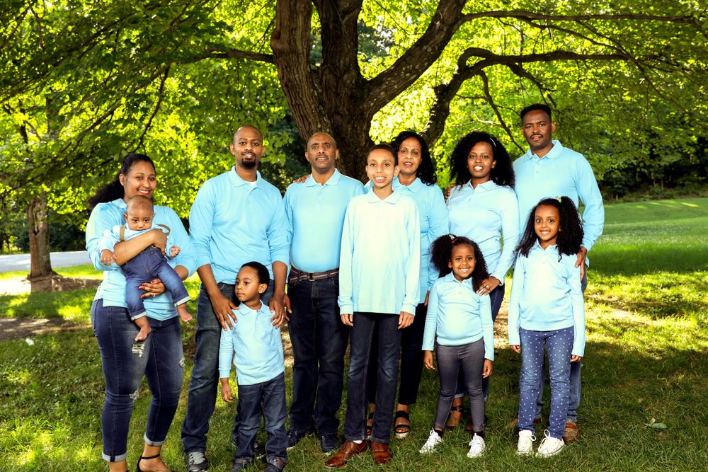 Extended family members in coordinating light blue and white outfits pose for a group photo under a leafy tree.