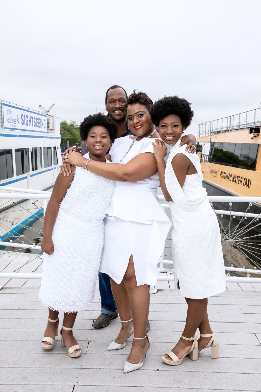 A family in matching white outfits embrace while posing on a dock with boats behind them.