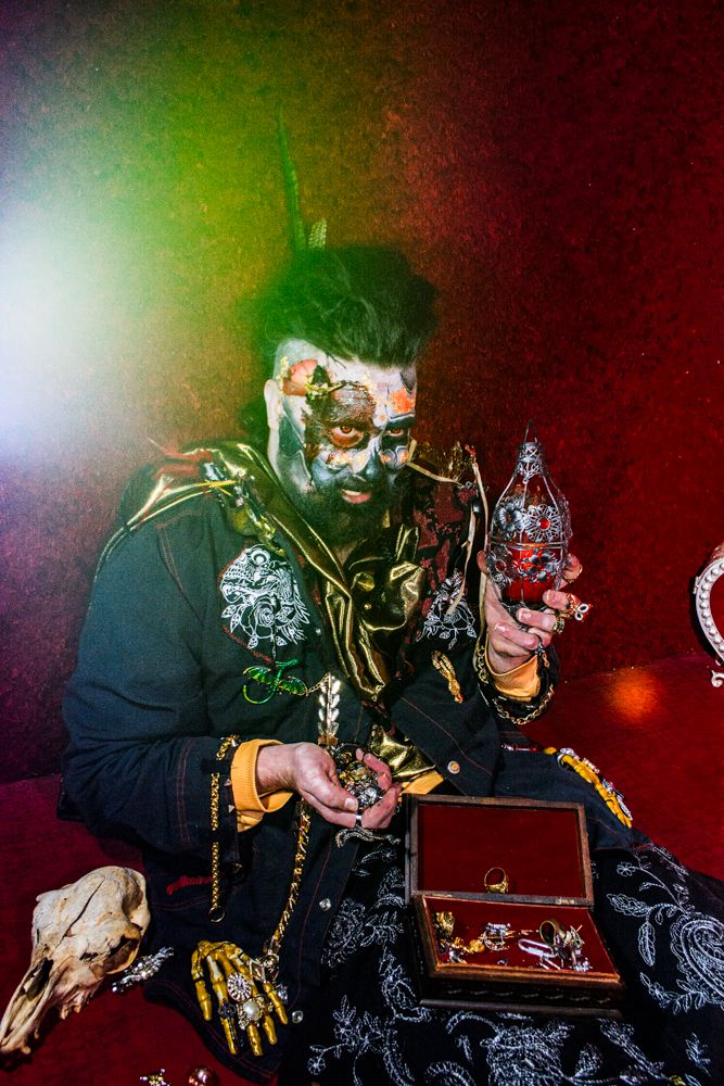 Gothic figure with dramatic makeup examines jewelry box with props and green lighting effects.