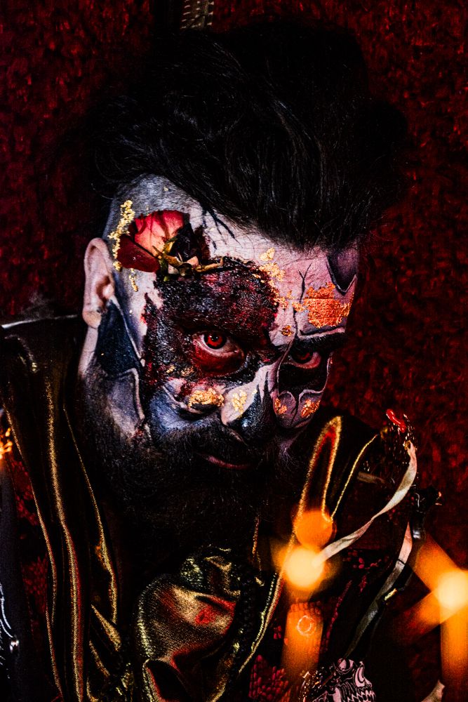 Dramatic close-up portrait with face paint and dark styling against red background.