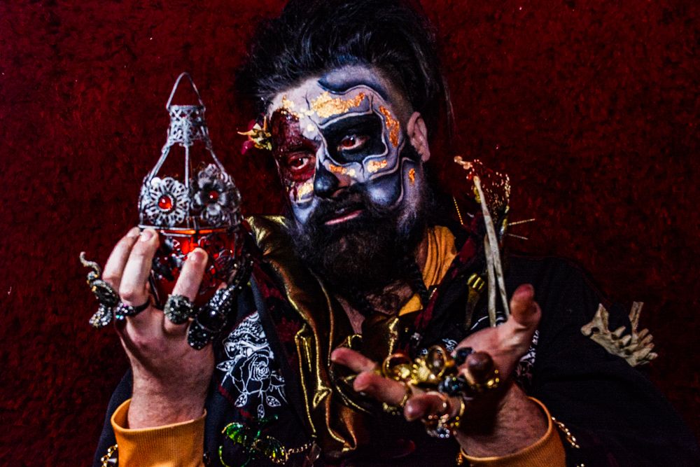 Gothic figure in dark clothing shows off steampunk accessories against red backdrop.