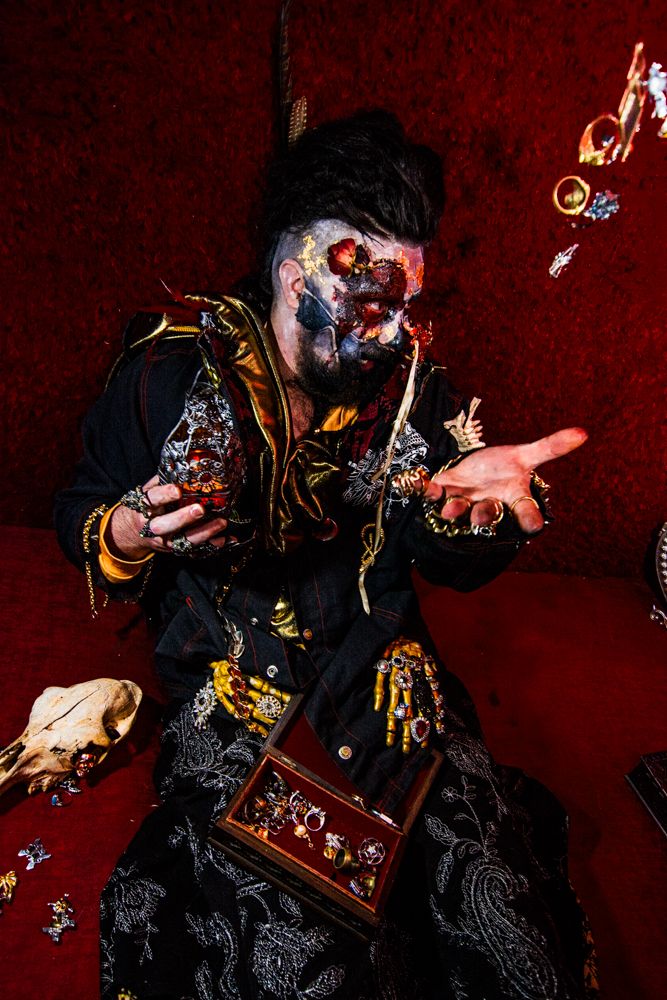 Dark-clad figure with face paint gestures dramatically with floating mechanical objects.