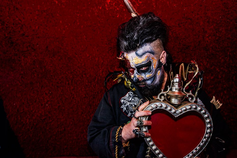 Figure in dark attire poses with decorative heart-shaped prop against red velvet background.