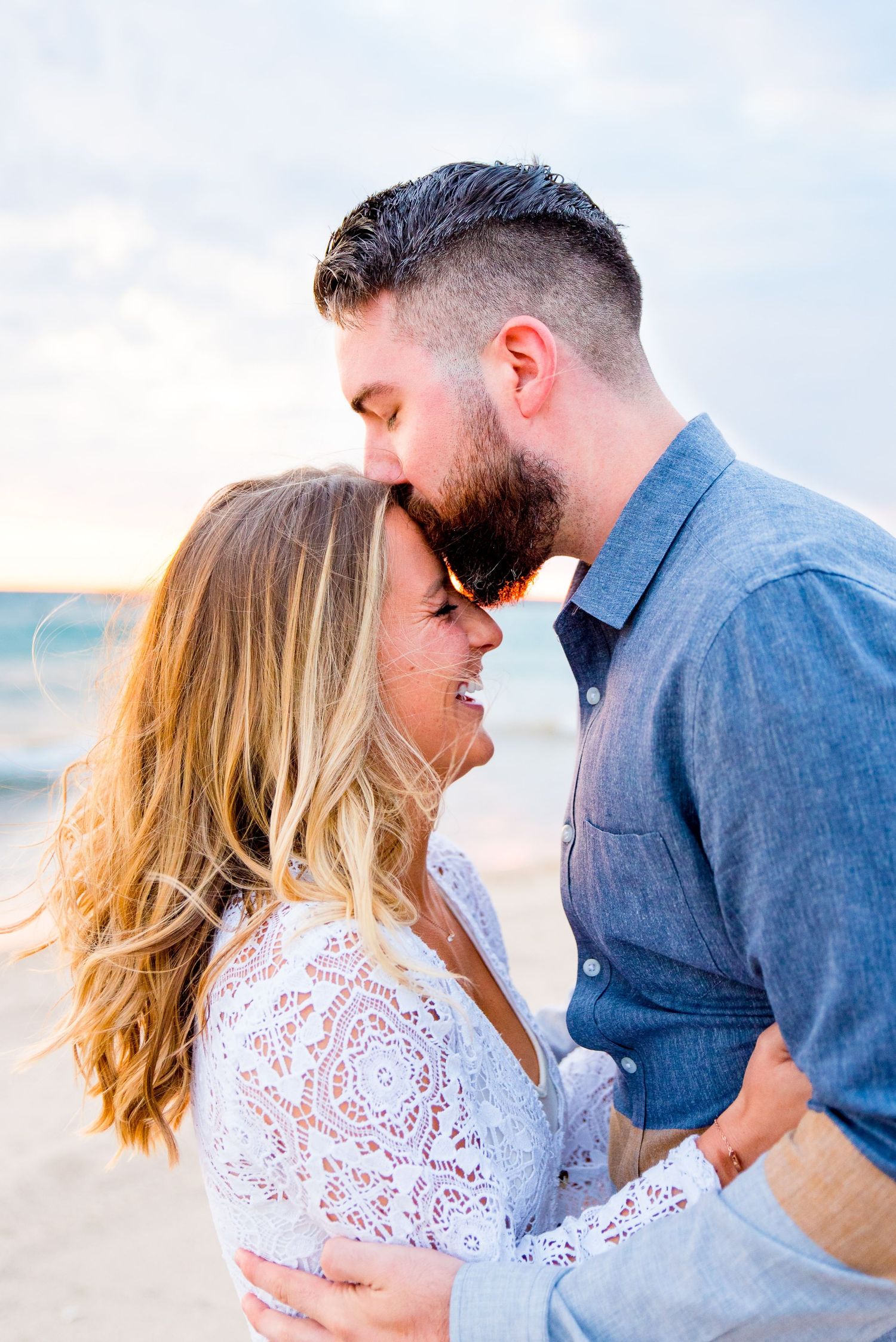 dark haired man kisses blonde woman on the forehead while she laughs in the wind