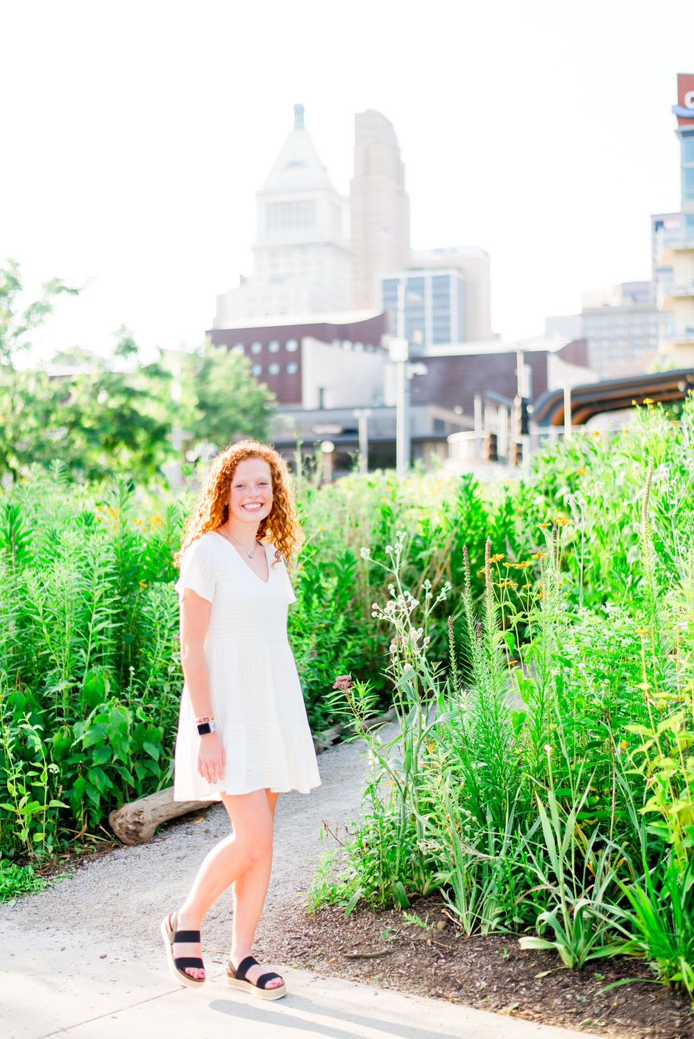 woman with red curly hair in white dress walks through flowers at Smale Park in downtown Cincinnati for senior pictures