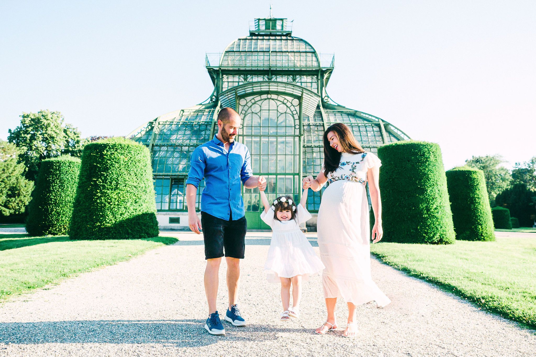Pregnant mother and father swinging their dauther in a bright sunny day at the Schönbrunn Palmenhaus in Vienna