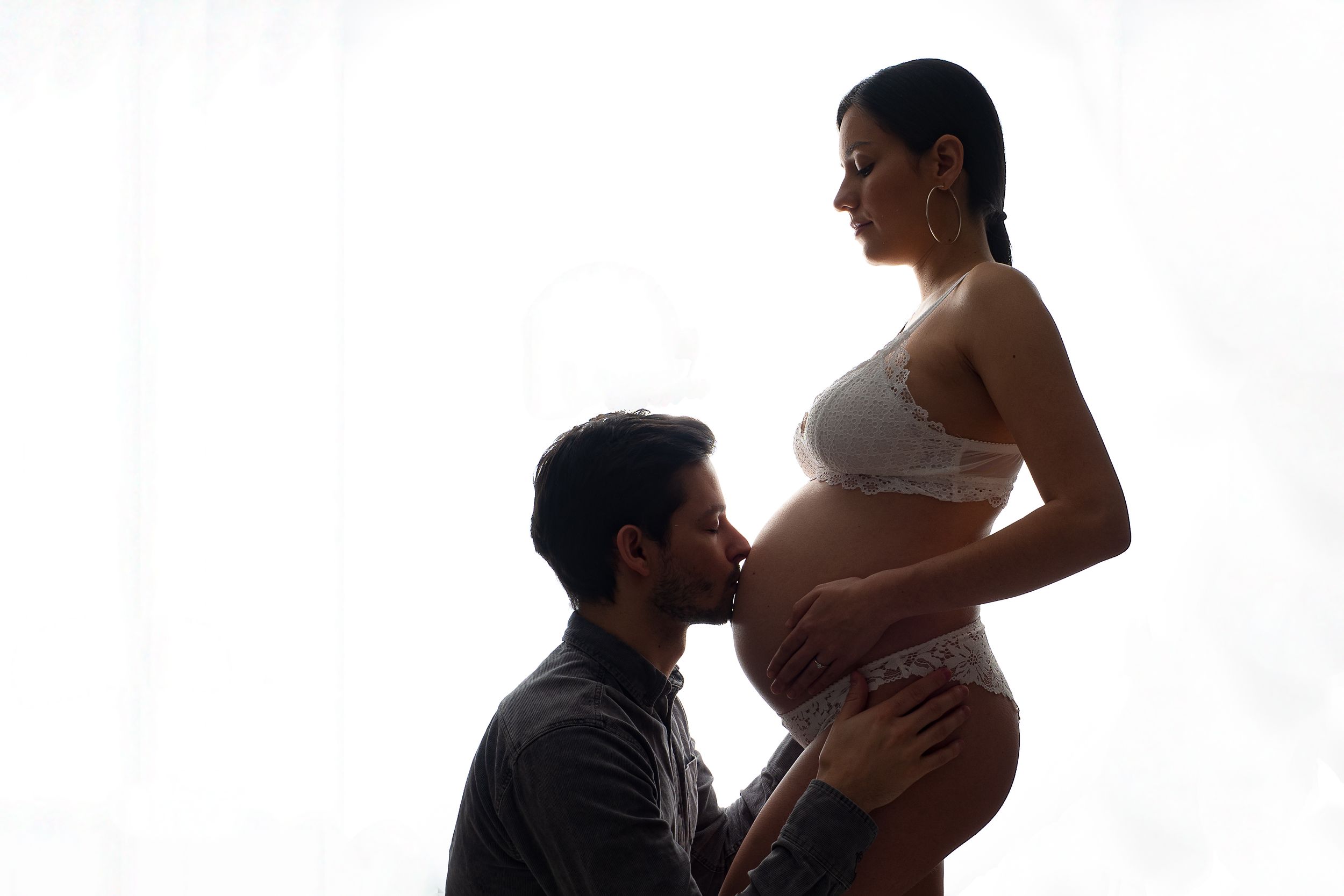 Pregnant woman and man kissing her belly with white underwear in photo studio Mödling
