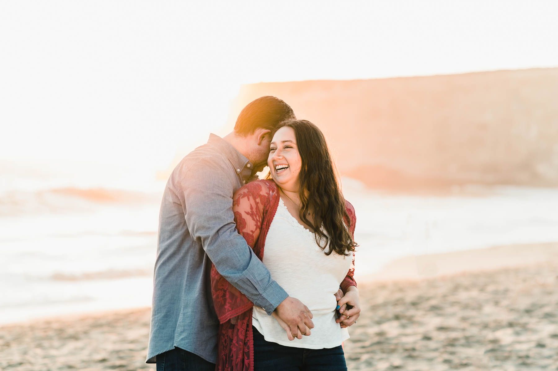 Couple in love sharing a laugh on the beach with the sun setting behind them.