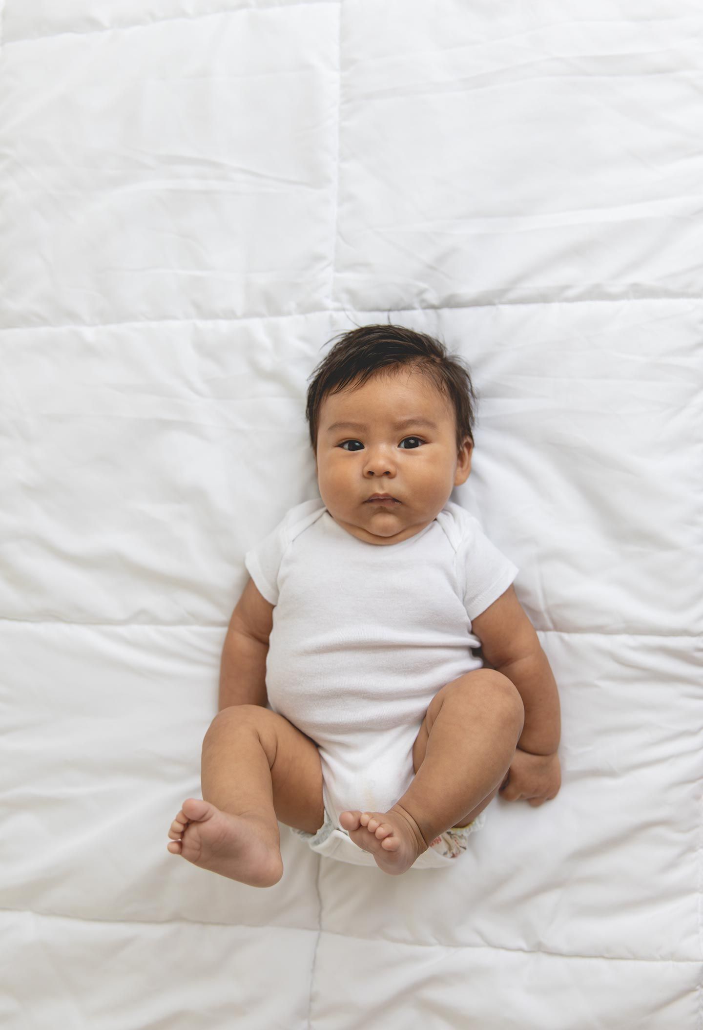 3 month old baby laying on white bedding and in a white onesie looking at the camera with a serious face.