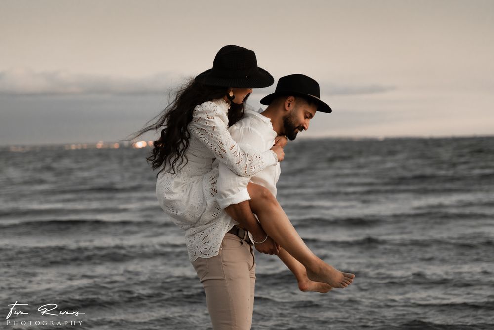 Playful beachside moment with couple in white outfits and black hats against ocean backdrop.