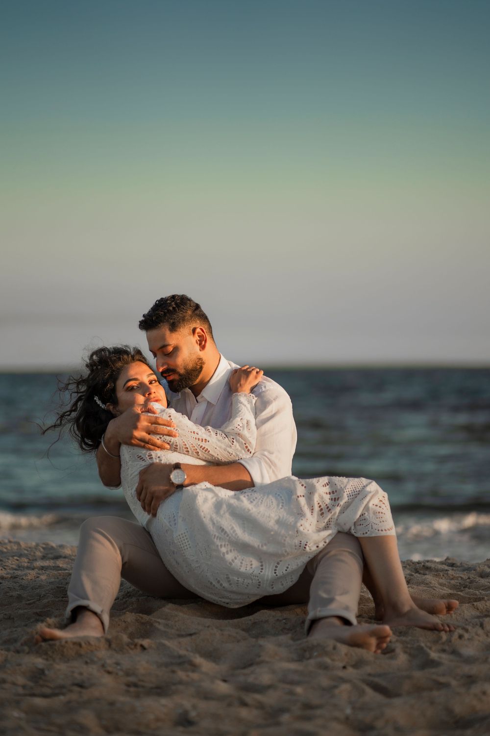 Couple in white clothing embraces romantically while sitting on sandy beach at sunset.