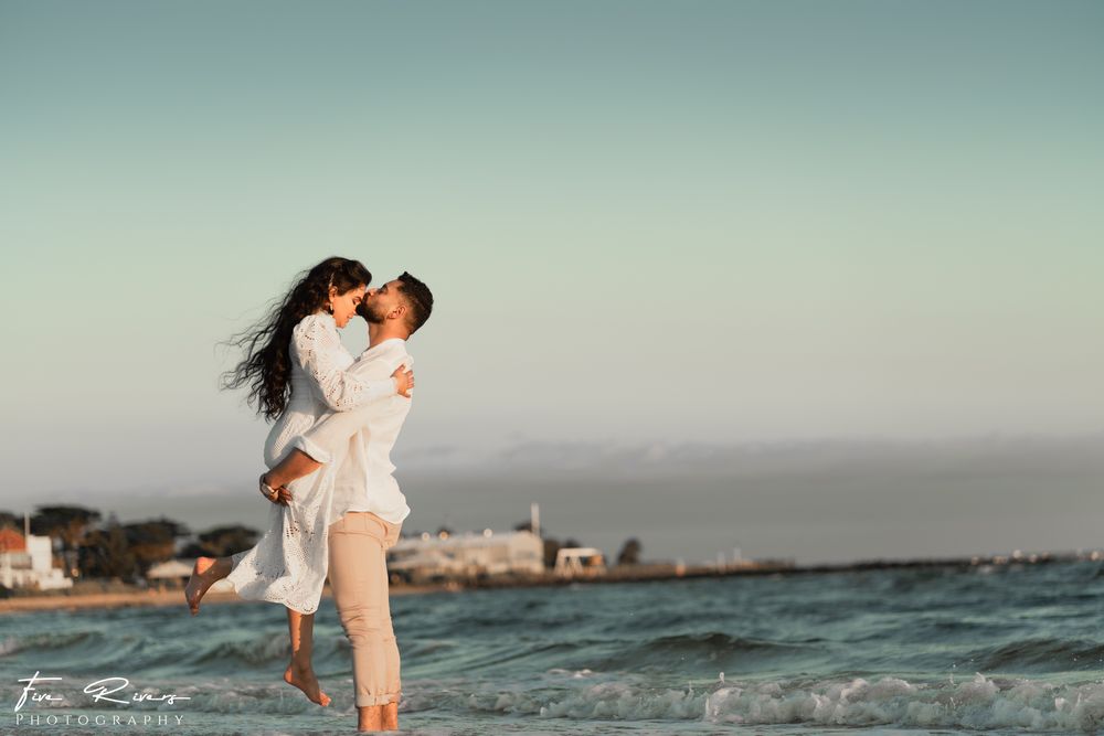 Romantic couple embracing on beach at sunset with ocean waves and coastal buildings in background.