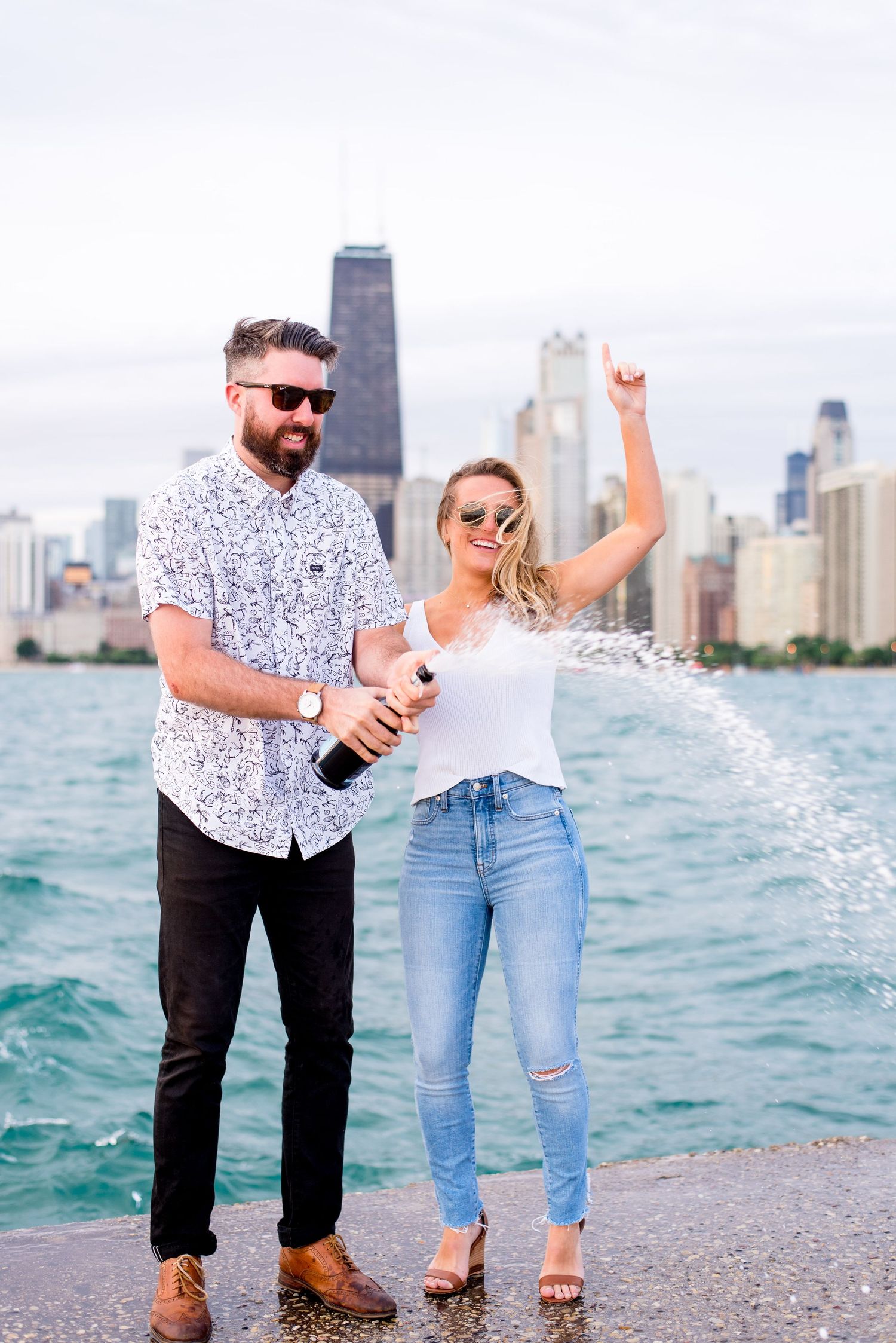 man and woman in sunglasses pop champagne at Chicago North Avenue Beach to celebrate their engagement