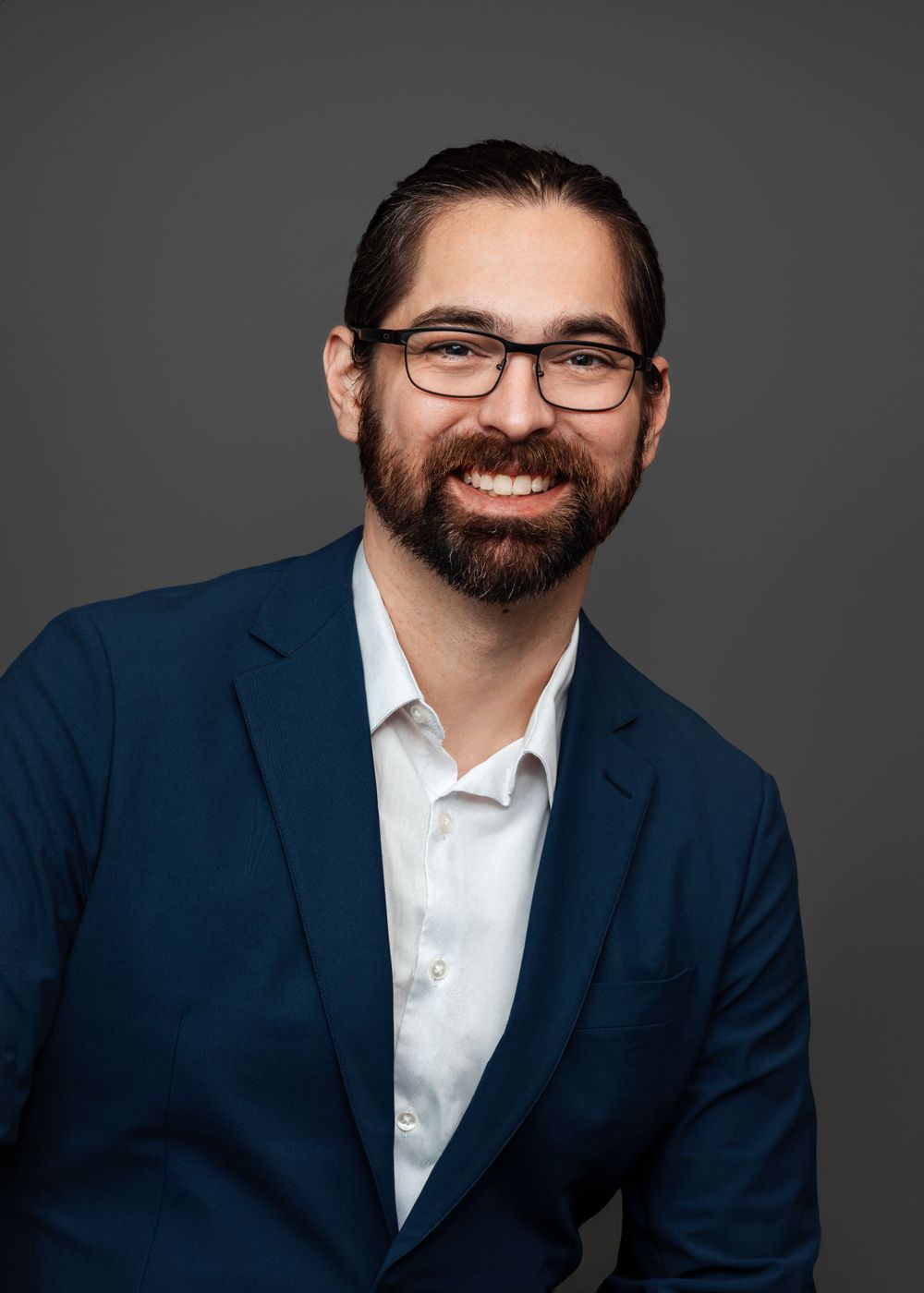 Professional headshot of a bearded man in glasses and navy blazer against gray background.