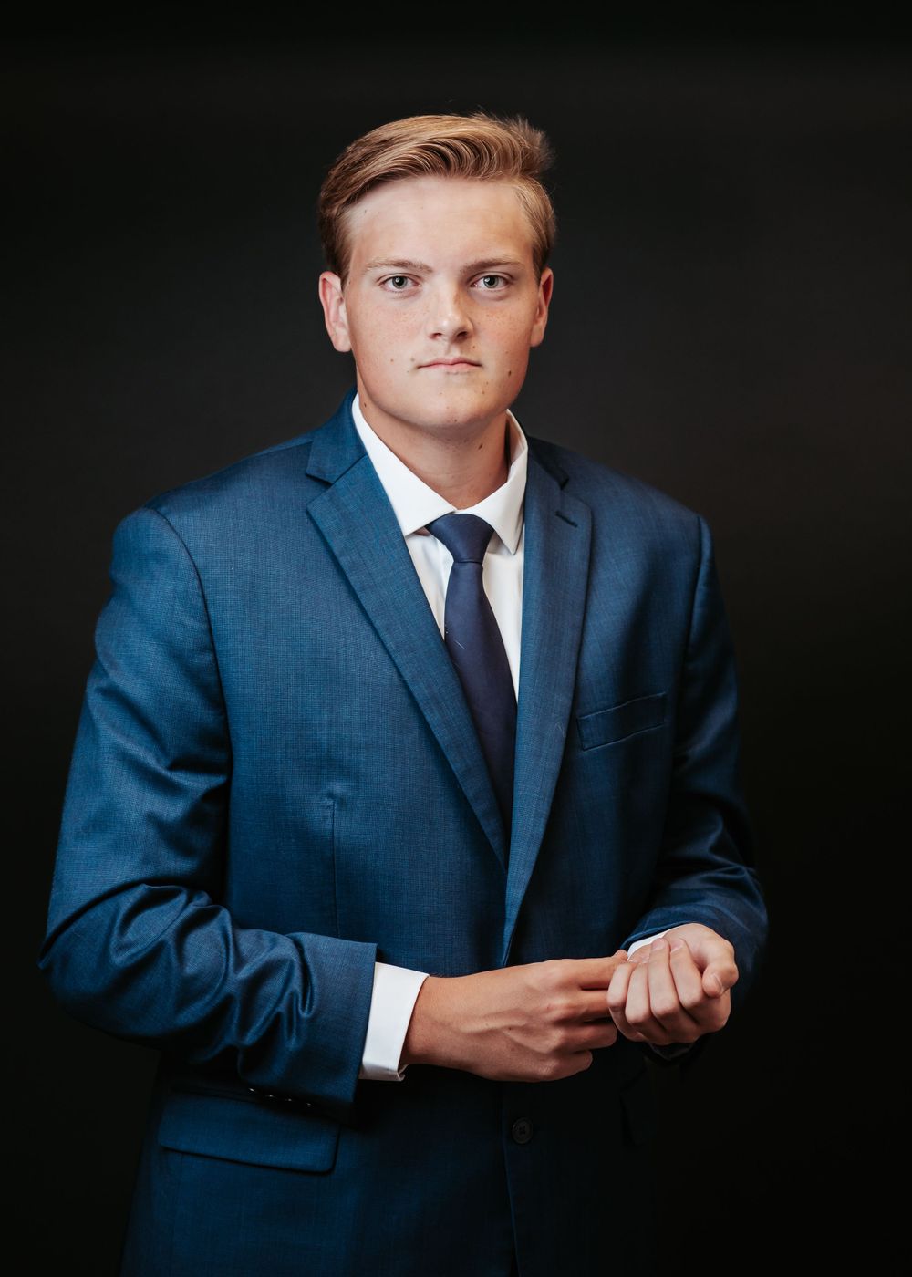 Professional headshot of a person in a navy blue suit and tie against a dark background.