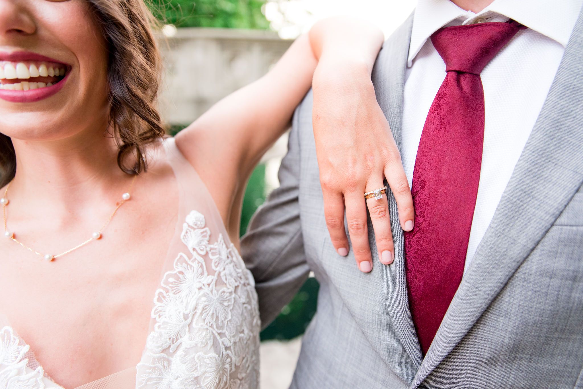 bride smiling with pink lipstick, resting elbow on groom's shoulder showing off her jewelry, his tie, and their clothing