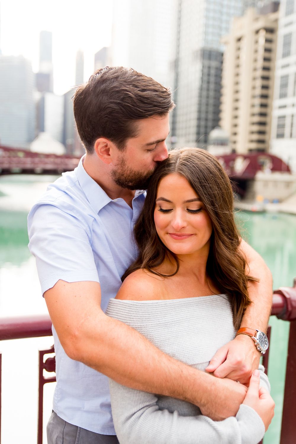 man with brown hair hugging fiance from behind kissing her by Chicago River for sunrise engagement pictures in Chicago