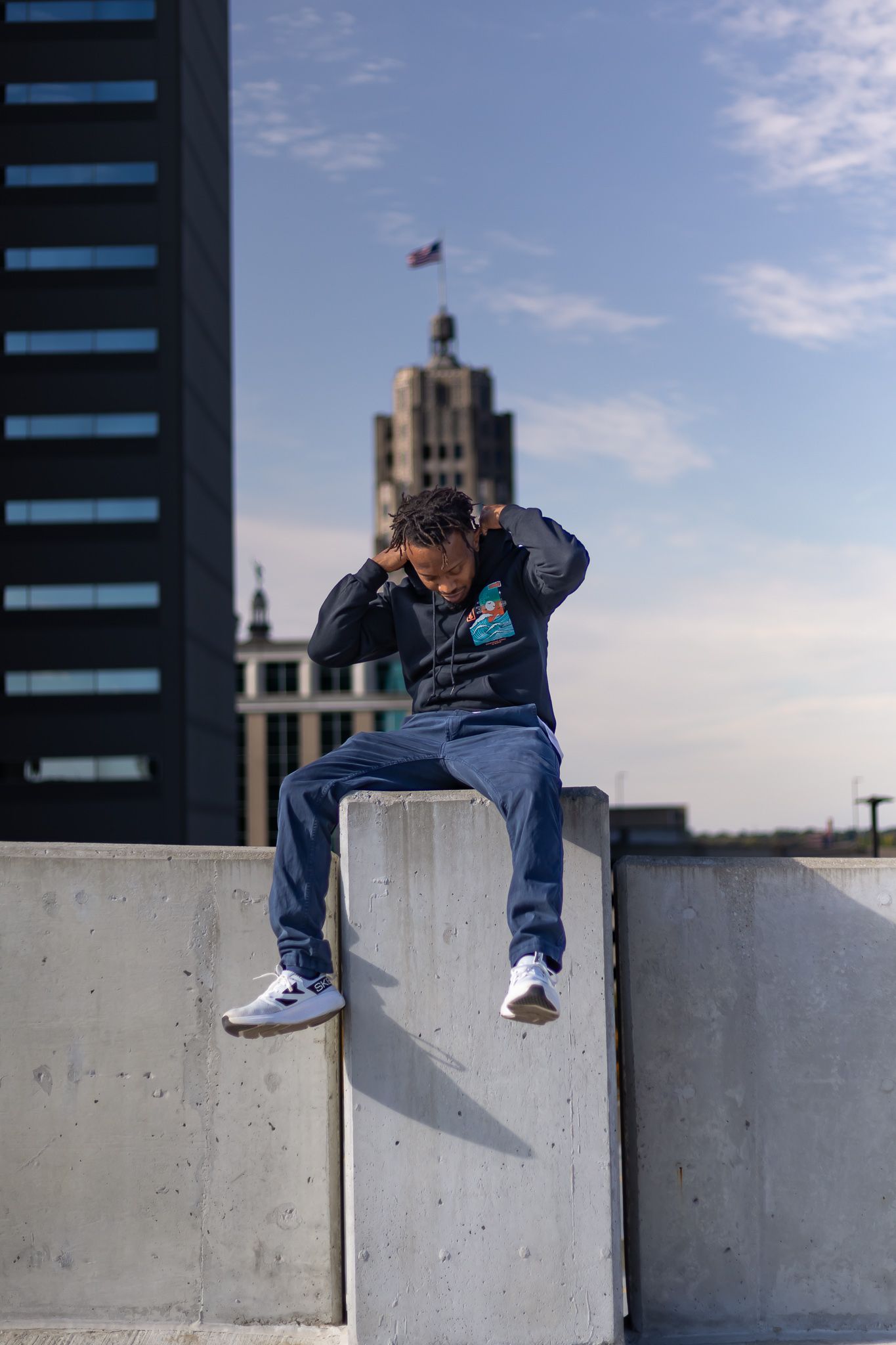 Young person sitting on a concrete wall with city buildings and flag in the background.